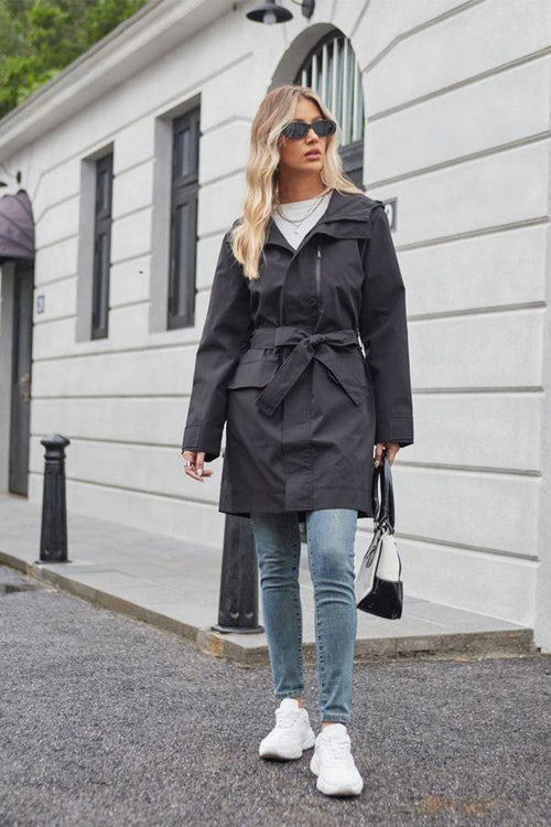 Woman wearing a black water-resistant windbreaker trench jacket with belt, jeans, white sneakers, and sunglasses walking on city street