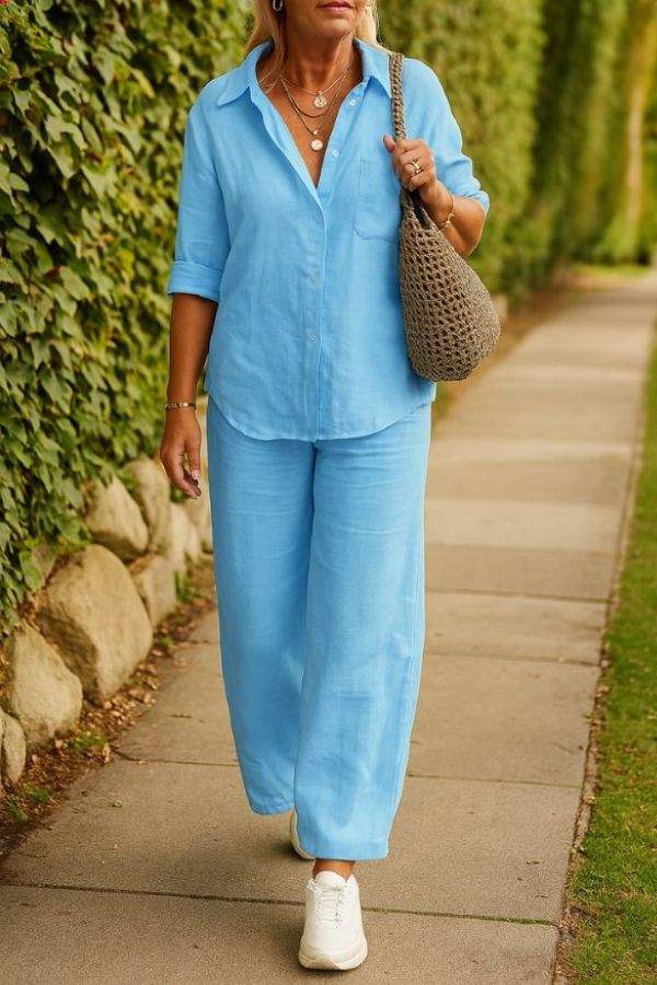 Woman wearing light blue relaxed shirt and trousers two-piece set with white sneakers and a woven handbag outdoors on a pathway