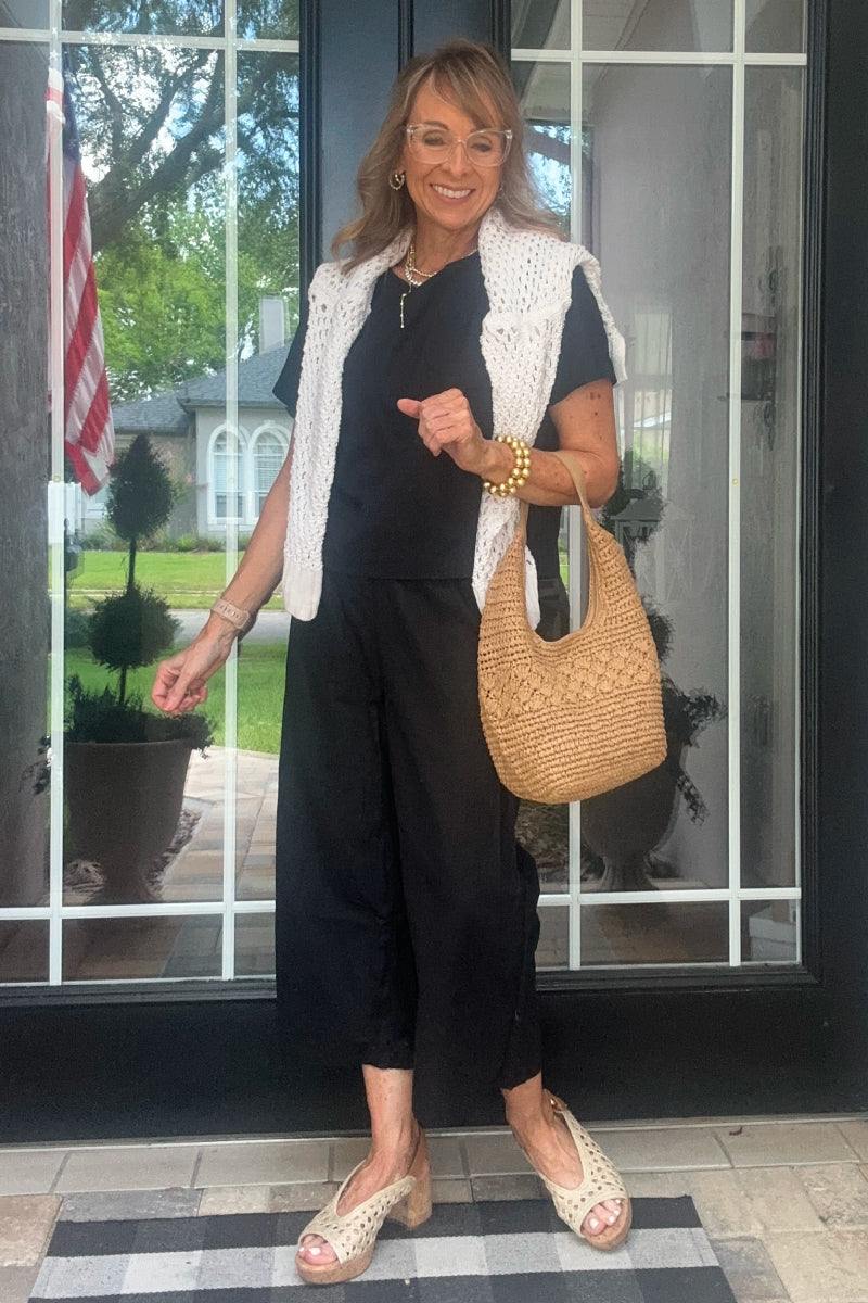 Woman wearing black loose fit co-ord set with wide-leg pants and short-sleeve top, holding woven handbag, white knit vest draped over shoulders.