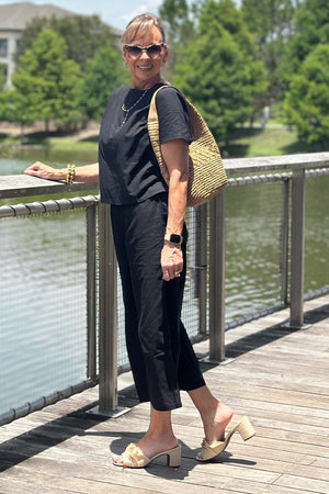 woman wearing black loose fit co-ord set with short sleeves and wide-leg pants standing on boardwalk with sunglasses and straw bag