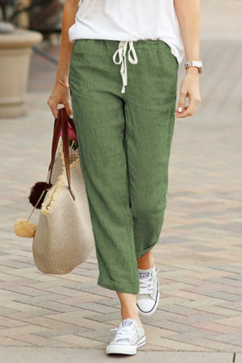 Woman wearing green relaxed fit straight pants with white drawstring and white sneakers walking on paved street carrying beige tote bag