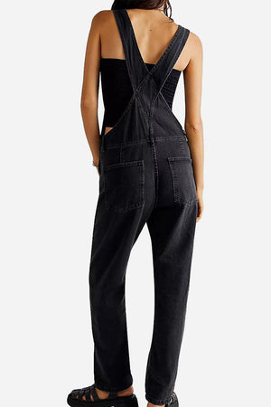 Back view of woman wearing black denim-look tapered-leg adjustable bib overalls with cross-back straps and pockets