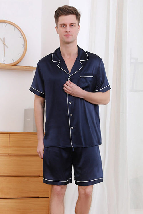 Man wearing navy blue Mulberry silk couples pajamas with white piping details in a bright room.