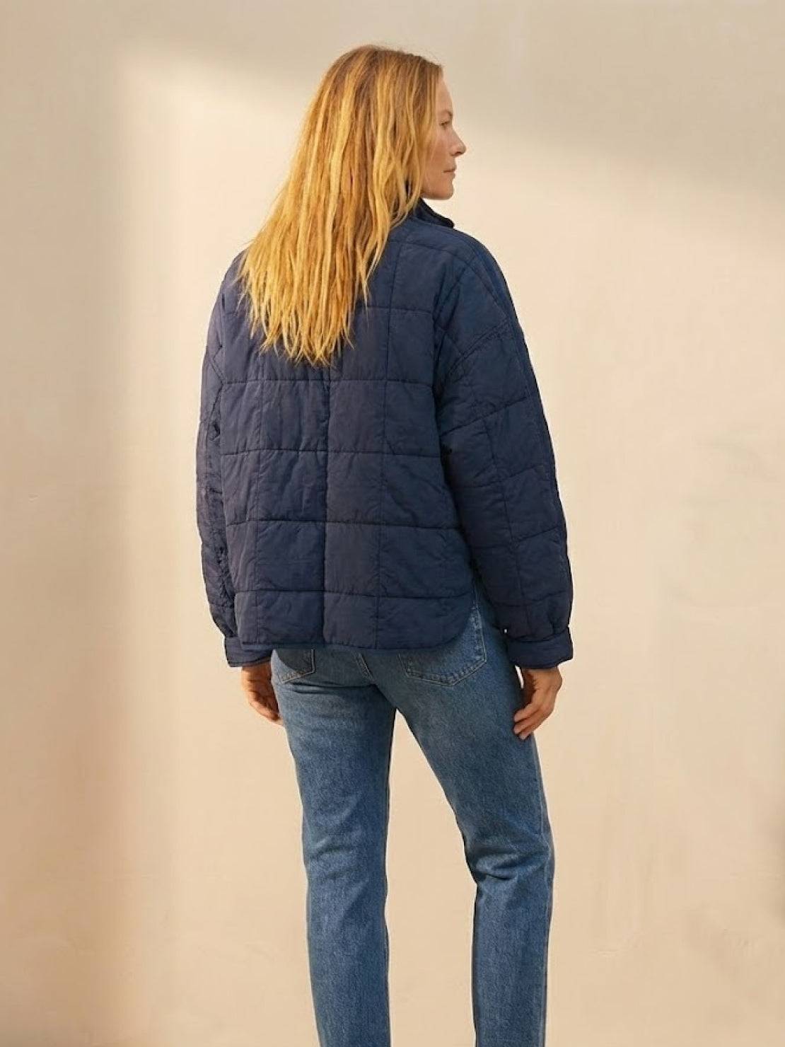 woman wearing navy blue lightweight puffer travel jacket and jeans standing against plain background