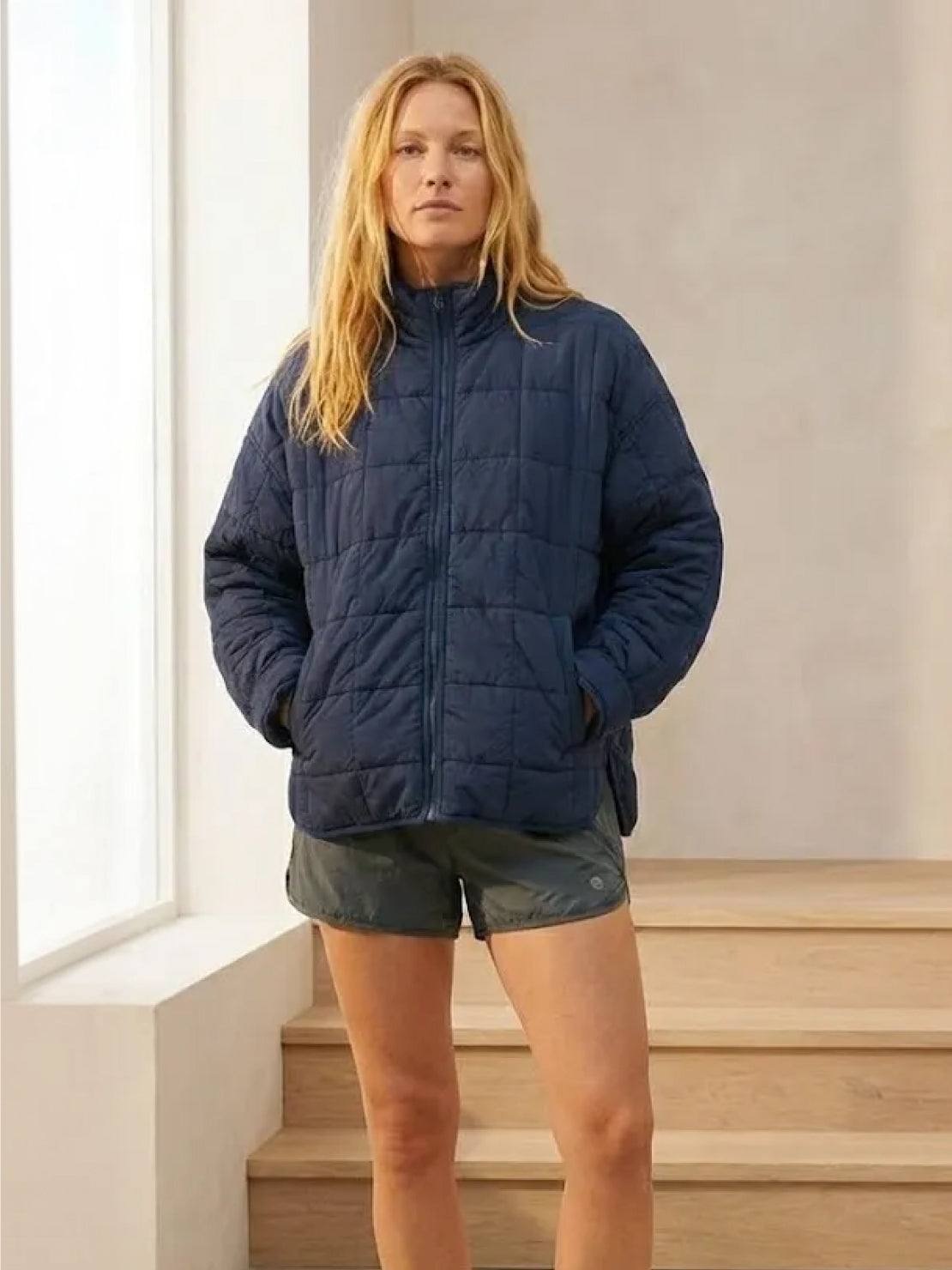 Woman wearing a navy lightweight puffer travel jacket with shorts indoors.