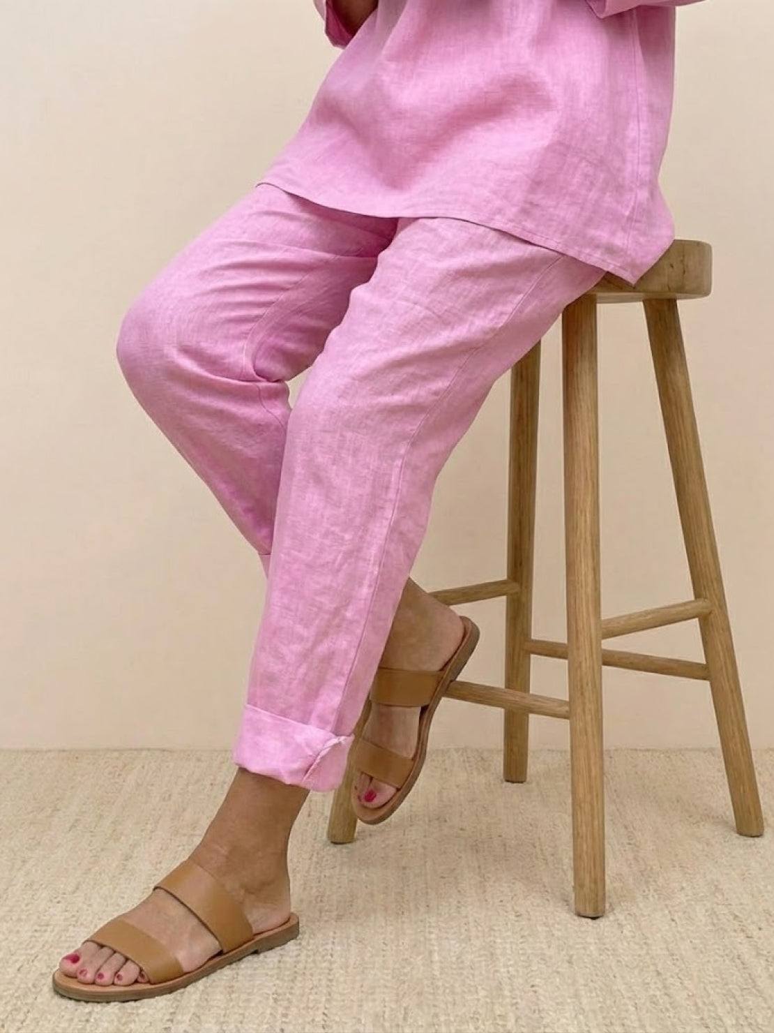 Pink asymmetrical two-piece trouser set with rolled-up sleeves and tapered pants paired with tan sandals sitting on wooden stool