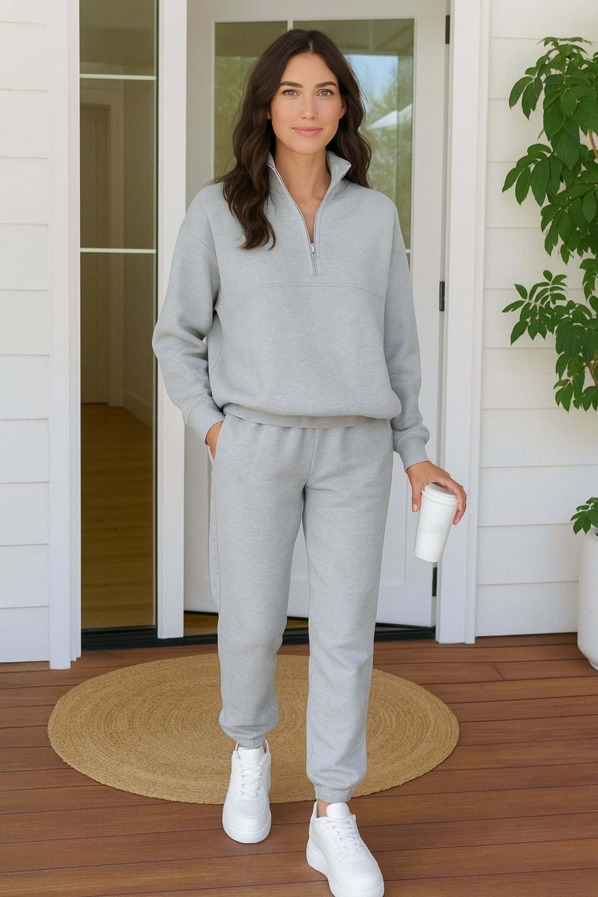 Woman wearing light gray cotton blend tracksuit with zip neckline and cropped hem, standing on wooden porch with white sneakers and holding a coffee cup