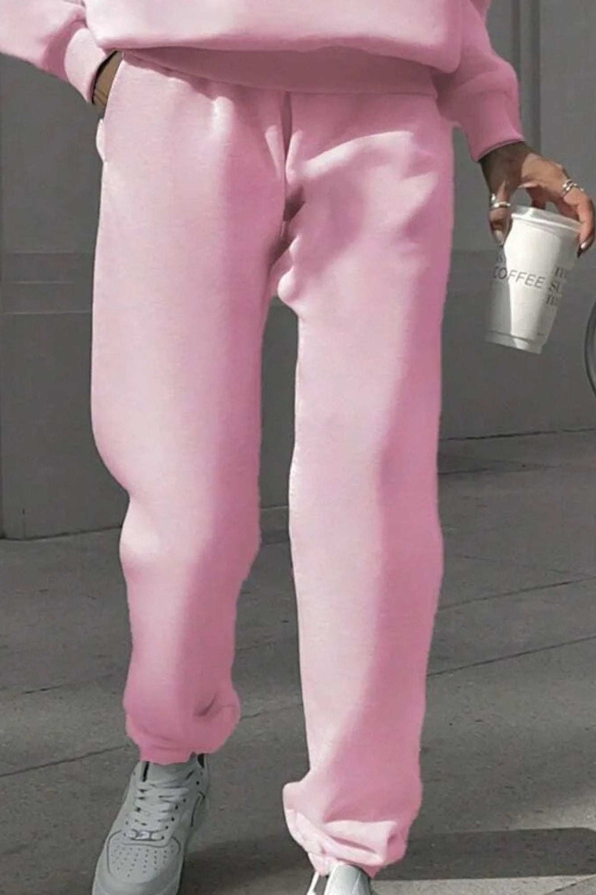 Close-up of a person wearing pink cotton blend tracksuit pants and gray sneakers holding a coffee cup outdoors