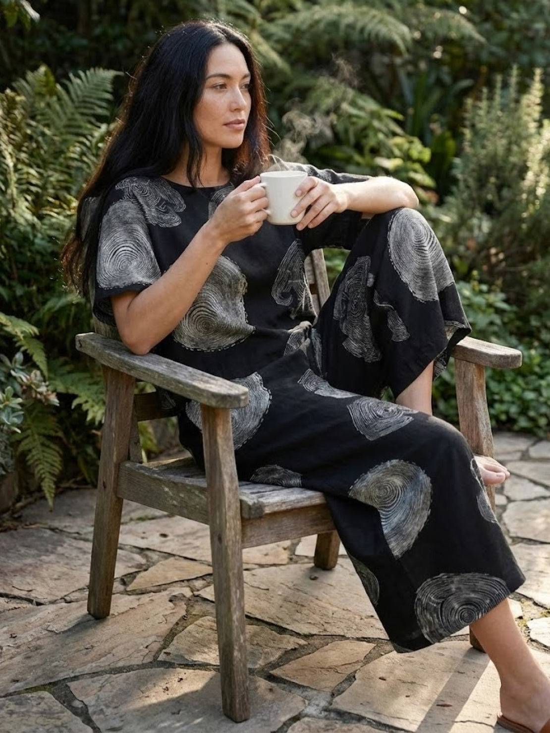 Woman wearing loose fit co-ord set with floral prints sitting on wooden chair holding a cup outdoors