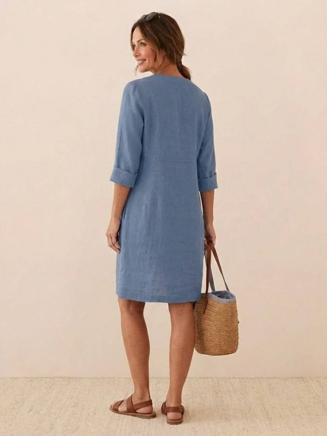 Woman wearing a casual blue cotton dress with rolled ¾ sleeves, standing and holding a woven handbag.