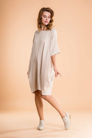 Woman wearing a nude oversized premium linen ovoid dress with short sleeves and discreet pockets, standing against a beige background