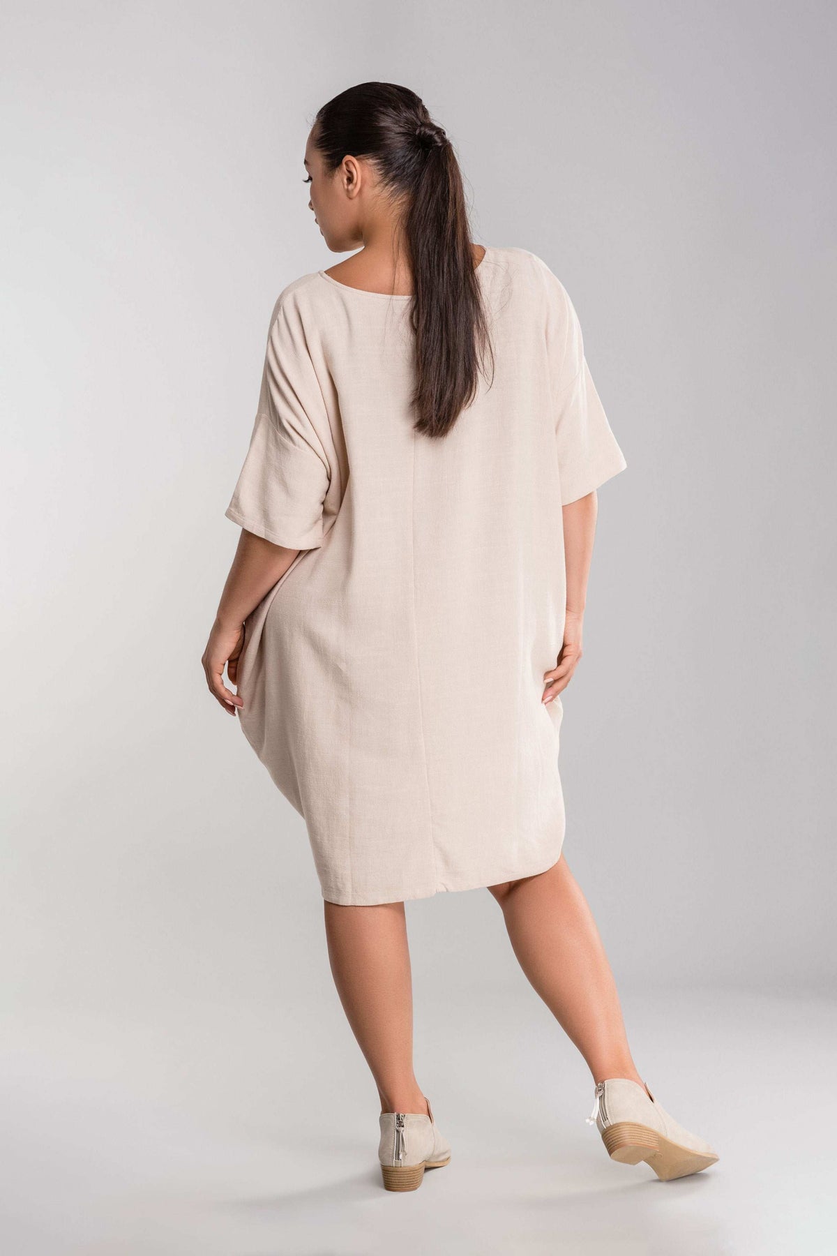 Back view of a woman wearing nude oversized premium linen ovoid dress with short sleeves and discreet pockets