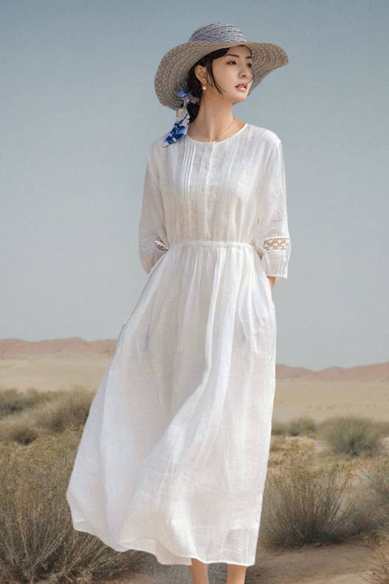 Woman wearing a white 100% linen coastal shift dress with long sleeves and floral lace details, styled with a wide-brimmed hat in a desert landscape.