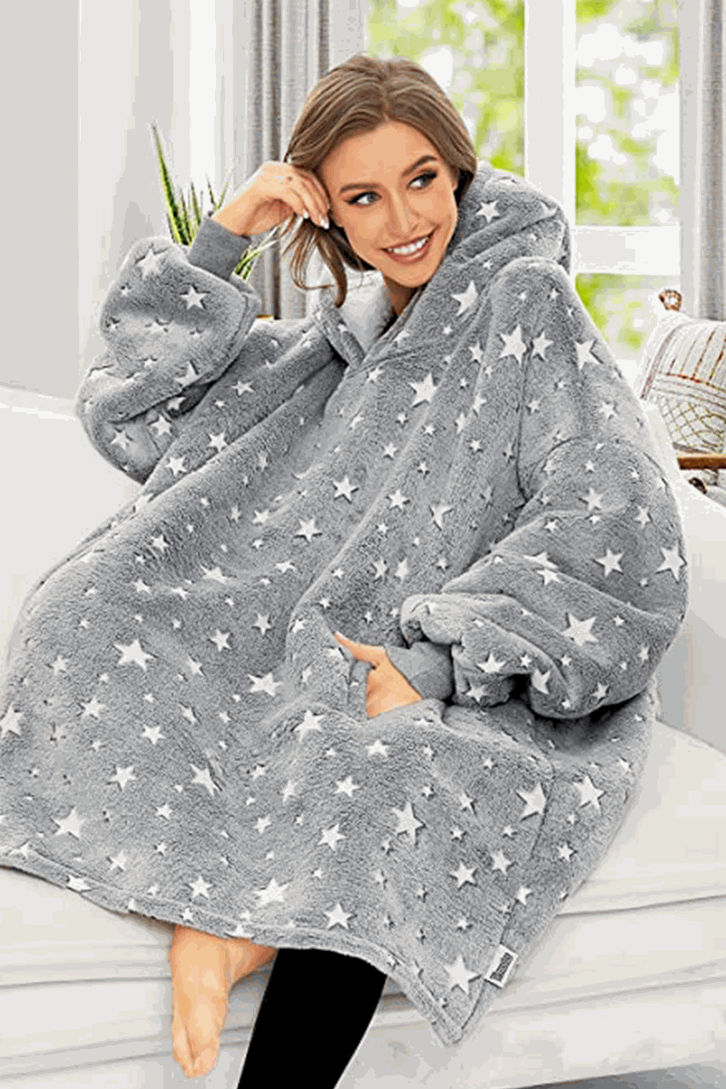 Woman wearing oversized gray Lazy Fleece Blanket Sweater with white stars, sitting indoors and smiling