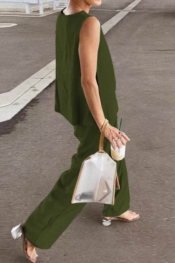 Woman wearing loose solid color sleeveless two-piece set in olive green walking outdoors with transparent handbag and drink in hand