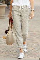 Woman wearing beige relaxed fit straight pants with drawstring waist and white sneakers walking outdoors carrying a woven tote bag