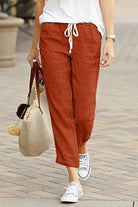 Woman wearing rust-colored relaxed fit straight pants with white drawstring and white sneakers walking outdoors carrying a beige tote bag