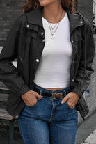 Woman wearing black outdoor hooded raincoat with white shirt and blue jeans standing against a brick wall