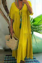 Yellow V-neck two-tiered maxi swing dress with loose sleeves and tiered flowing skirt, styled with a turquoise necklace and woven handbag.