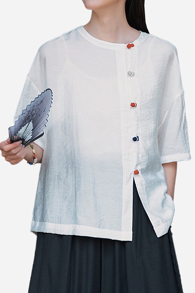 White regenerated cellulose blouse with asymmetrical colorful buttons, oversized fit, and short sleeves, styled with dark pants and holding a fan.