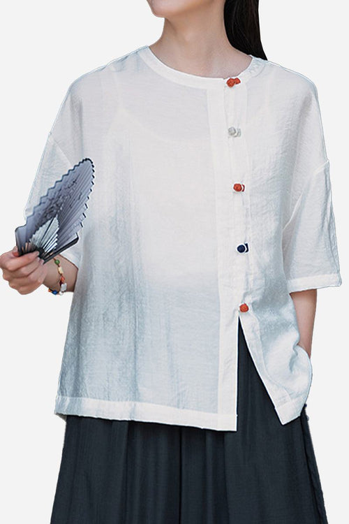 White regenerated cellulose blouse with asymmetrical colorful buttons, oversized fit, and short sleeves, styled with dark pants and holding a fan.