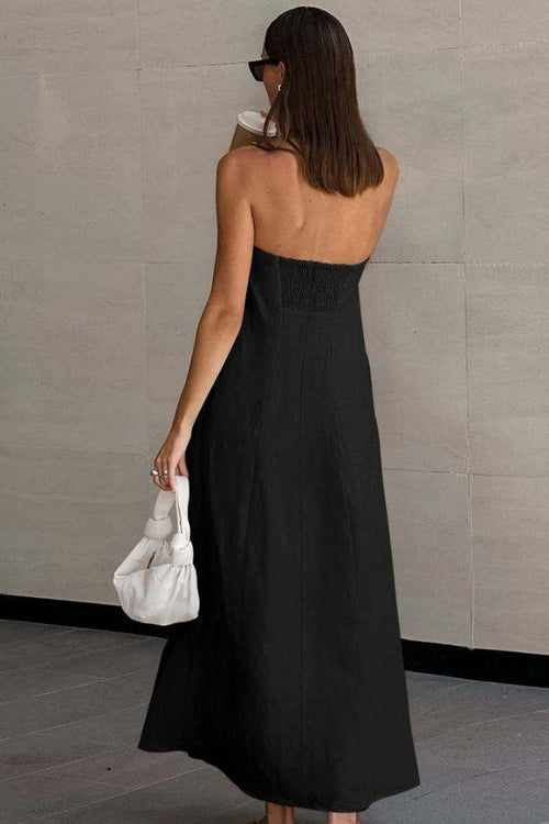 Back view of woman wearing black strapless cotton linen blend long dress with elastic back panel holding white handbag