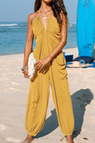 Woman wearing mustard yellow halter-neck wide-leg jumpsuit with elastic ankle cuffs on the beach holding white clutch bag Sunshine On Sand style