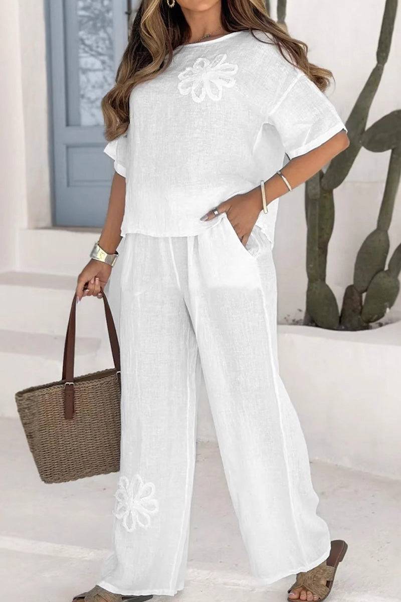 White flowy two piece set with floral embroidery, short sleeves, and wide leg pants, styled with a woven tote bag and casual sandals.