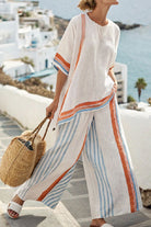 Woman wearing Santorini Sunset Striped two-piece set with white, orange, and blue stripes, walking outdoors near white buildings and sea with a woven bag and sandals