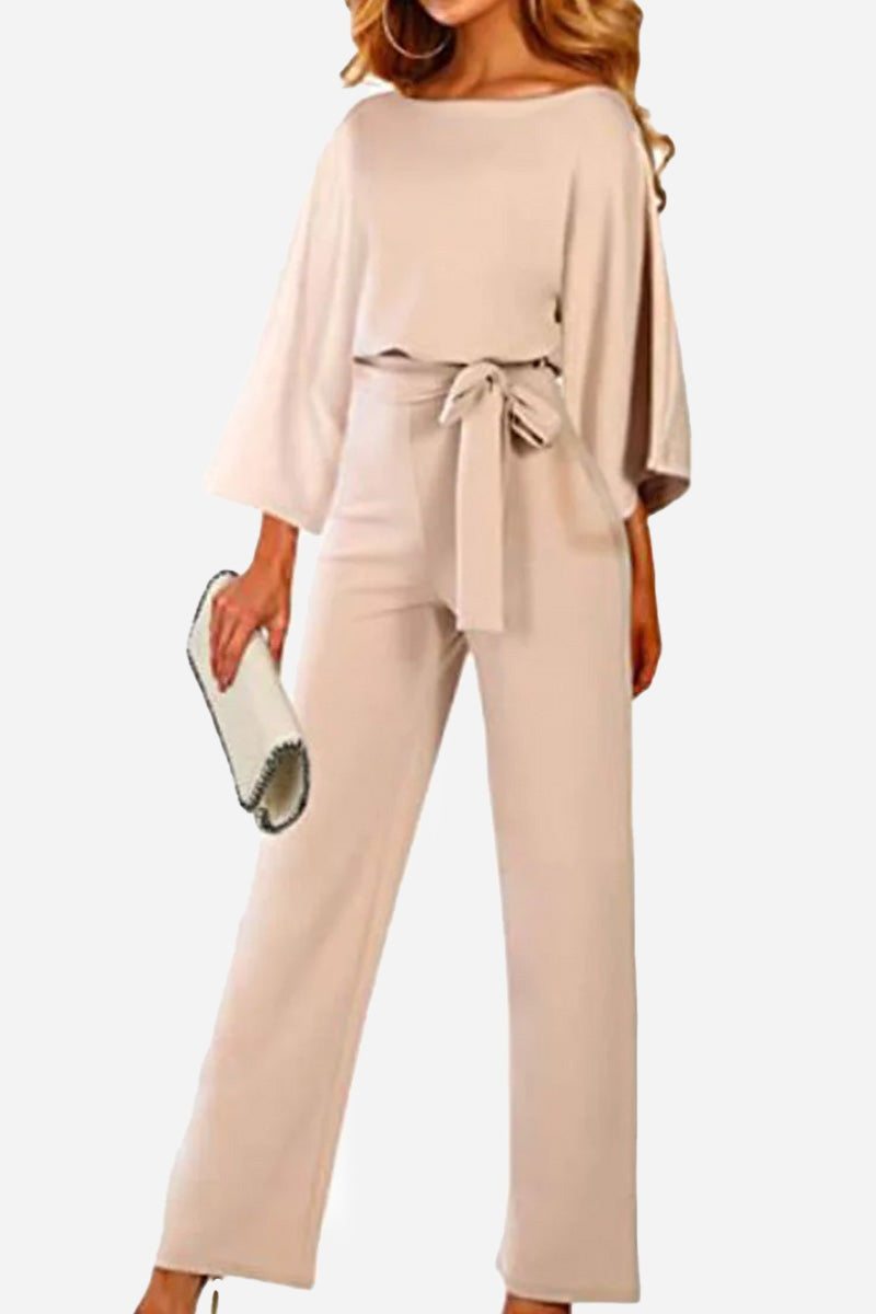 Woman wearing beige casual long sleeve belted jumpsuit with wide sleeves and round neck, holding a white clutch bag.