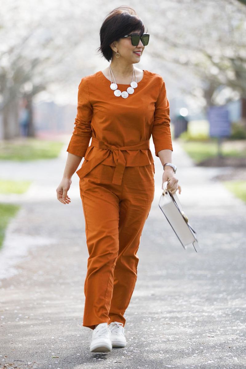 Woman wearing rust-colored cotton-linen monochrome relaxed longline top and wide-leg pants walking outdoors with sunglasses and white sneakers