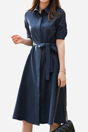 Woman wearing a navy radiance belted shirt dress with short sleeves and a detachable waist belt, holding a black handbag.