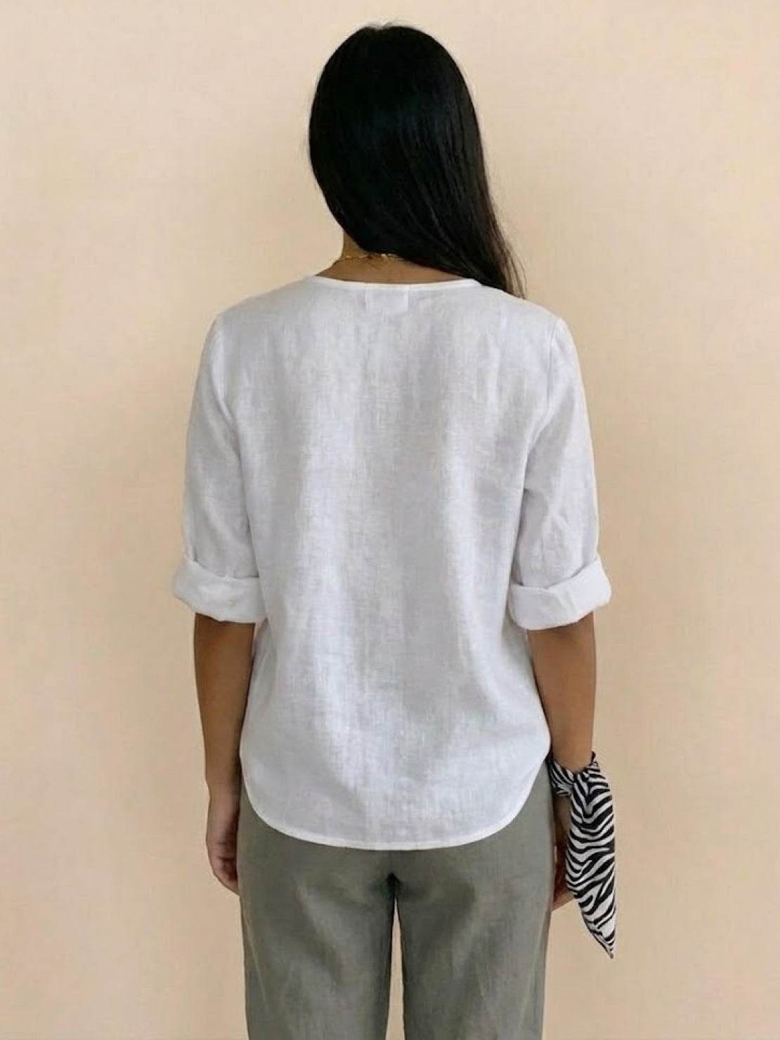 Back view of a woman wearing a soft white pure linen age-defying button detail shirt with rolled sleeves and olive pants, holding a patterned scarf.