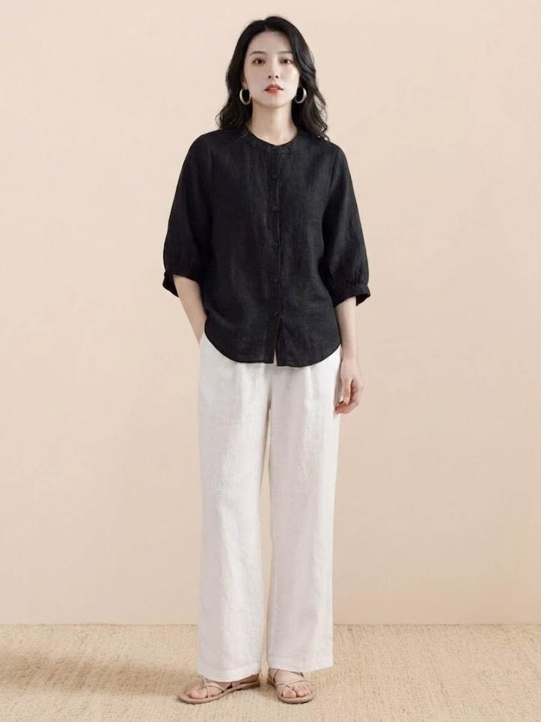 Woman wearing relaxed loose breathable linen black blouse and white pants standing against beige background