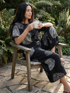Woman wearing loose fit co-ord set with floral prints sitting on wooden chair holding a cup outdoors