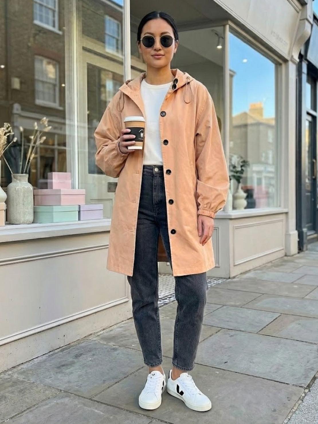 Woman wearing Rain Couture Windbreaker in light beige oversized water-resistant jacket with hood, black jeans, white sneakers, and sunglasses outdoors.