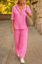 Woman wearing pink relaxed shirt and trousers two-piece set with white sneakers and a woven bag walking outdoors