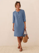 Woman wearing a knee-length blue cotton dress with half-button front and rolled ¾ sleeves holding a woven handbag.