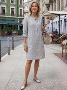 woman wearing gray cotton dress with rolled-up sleeves standing outdoors in city street with buildings and café seating in background