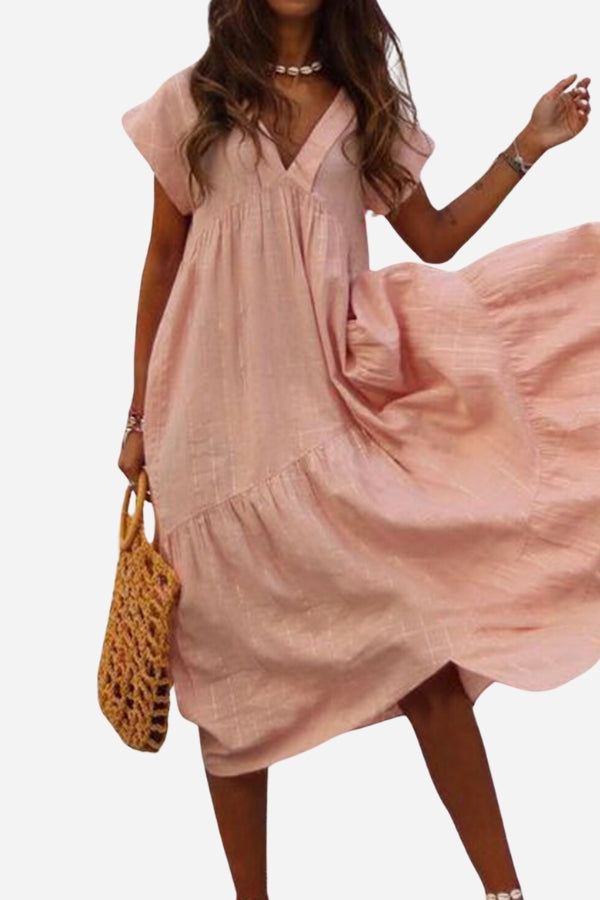 Woman wearing a pink V neck tiered cotton midi dress with short sleeves and frill hem holding a woven handbag